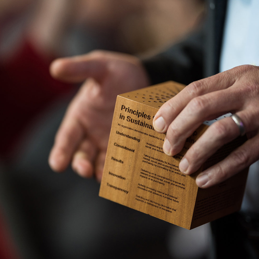 A hand presents an engraved wooden award cube listing sustainable purchasing principles.